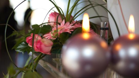 Blumenarrangement mit rosa Gerbera und Rosen, umgeben von dekorativen Kerzen auf einem Tisch.