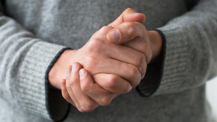 Nahaufnahme von gefalteten Händen einer Person in einem grauen Pullover.