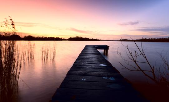 Holzsteg führt über ruhiges Wasser, umgeben von Gräsern und einem farbenprächtigen Sonnenuntergang.