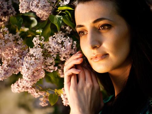 Nahaufnahme einer Frau mit langen, dunklen Haaren, die sanft von lila Fliederblüten umgeben ist.