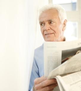 Älterer Mann mit weißem Haar liest eine aufgeschlagene Zeitung, während er an einem Fenster steht.